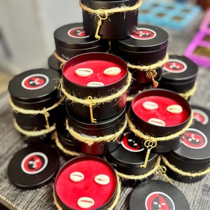 Puede incluir: Latas de velas negras con cera roja, adornadas con conchas marinas y envueltas con cuerda y llaves doradas. Las tapas presentan un gráfico rojo y negro. Una pila de velas se exhibe sobre una superficie de madera.