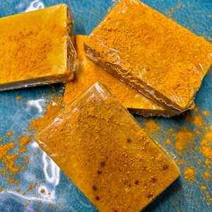 May include: Three bars of yellow soap with yellow powder sprinkled on top. The soap is wrapped in clear plastic and is sitting on a blue and white textured surface.