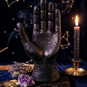 May include: A matte black palmistry hand sculpture featuring an engraved eye at the center of the palm and various celestial symbols on the fingers and wrist. The hand is positioned upright on a square black base with a detailed etched design.