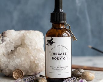 May include: A brown glass bottle of Hecate Ritual Body Oil with a black dropper and a key charm. The label features a witch silhouette and the words "Protection-Intuition-Transformation." The bottle is on a marble surface with crystals and lavender.