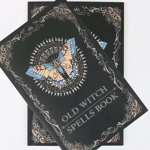 以下が含まれることがあります： 銀色の装飾的な縁取りと「OLD WITCH SPELLS BOOK」のテキストが付いた2冊の黒い呪文の本。表紙には、青、タン、白の蝶のイラストが、銀色のサンバーストのデザインの中に描かれています。