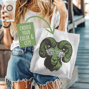 May include: A white tote bag with a green ram wearing sunglasses and the text "Rose Bowl Rams". The bag is being held by a person wearing a white t-shirt and denim shorts.