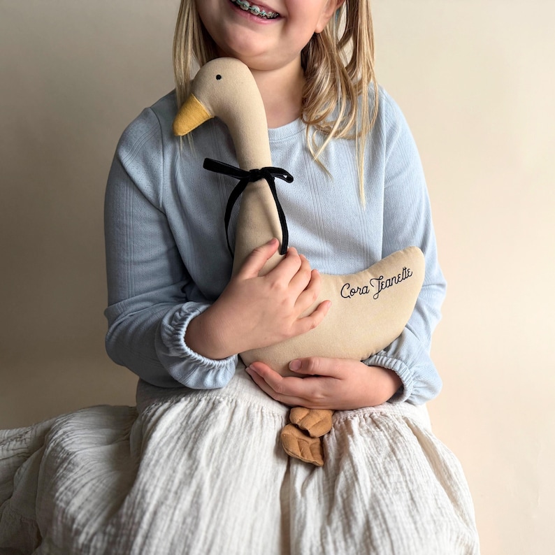 May include: A beige plush goose toy with a yellow beak and black ribbon around the neck. The goose has the name "Cora Jeanelle" embroidered on its side. The toy is held by a person wearing a light blue long-sleeved shirt and a white skirt.