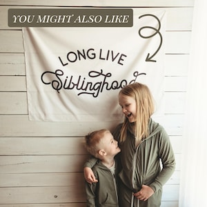 May include: A white banner with the text "LONG LIVE Siblinghood" in black script. Two children are wearing olive green zip-up hoodies. The banner is hanging on a white wooden wall.