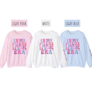 K&ouml;nnte beinhalten: Drei lang&auml;rmelige Sweatshirts in Hellrosa, Wei&szlig; und Hellblau mit dem Text "IN MY FIGURE SKATING ERA" in einer Retro-Schriftart mit rosa, lila und blauen Farben.