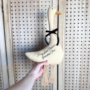 Puede incluir: Un peluche de ganso de tela color crema con cuello largo, pico beige y dos patas beige. Una cinta negra est&aacute; atada alrededor del cuello. El cuerpo tiene bordado el texto en negro, A hug from Heaven, Grandma Terri, en letra cursiva.