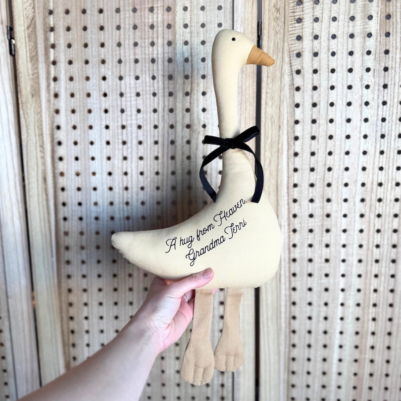 Puede incluir: Un peluche de ganso de tela color crema con cuello largo, pico beige y dos patas beige. Una cinta negra est&aacute; atada alrededor del cuello. El cuerpo tiene bordado el texto en negro, A hug from Heaven, Grandma Terri, en letra cursiva.
