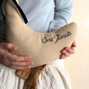 May include: A beige crescent-shaped pillow with the name "Cora Jeanette" embroidered in black script. The pillow has a black ribbon and a brown fabric detail. The pillow is held by a person wearing a light blue shirt and a white skirt.