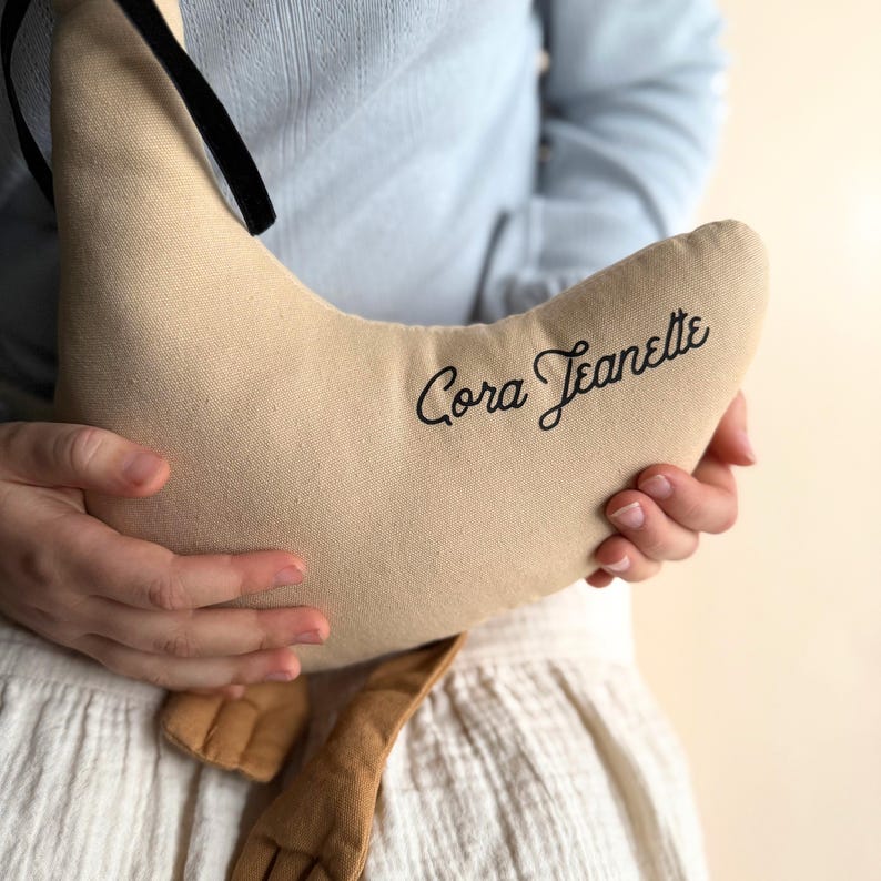 May include: A beige crescent-shaped pillow with the name "Cora Jeanette" embroidered in black script. The pillow has a black ribbon and a brown fabric detail. The pillow is held by a person wearing a light blue shirt and a white skirt.