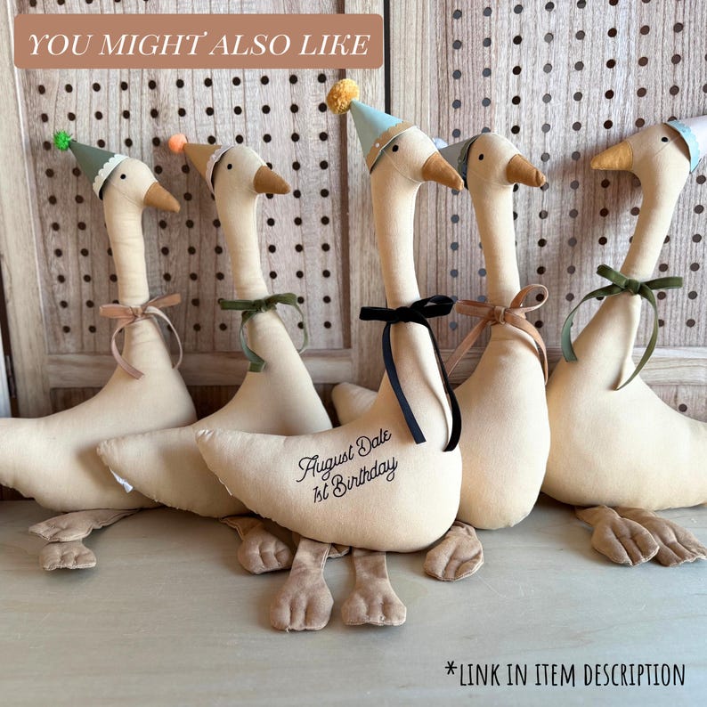 May include: Five plush geese with beige bodies, brown feet, and long necks. Each goose wears a party hat and a ribbon bow. One goose has the text "August Date 1st Birthday" embroidered on its body. The text "YOU MIGHT ALSO LIKE" is at the top.