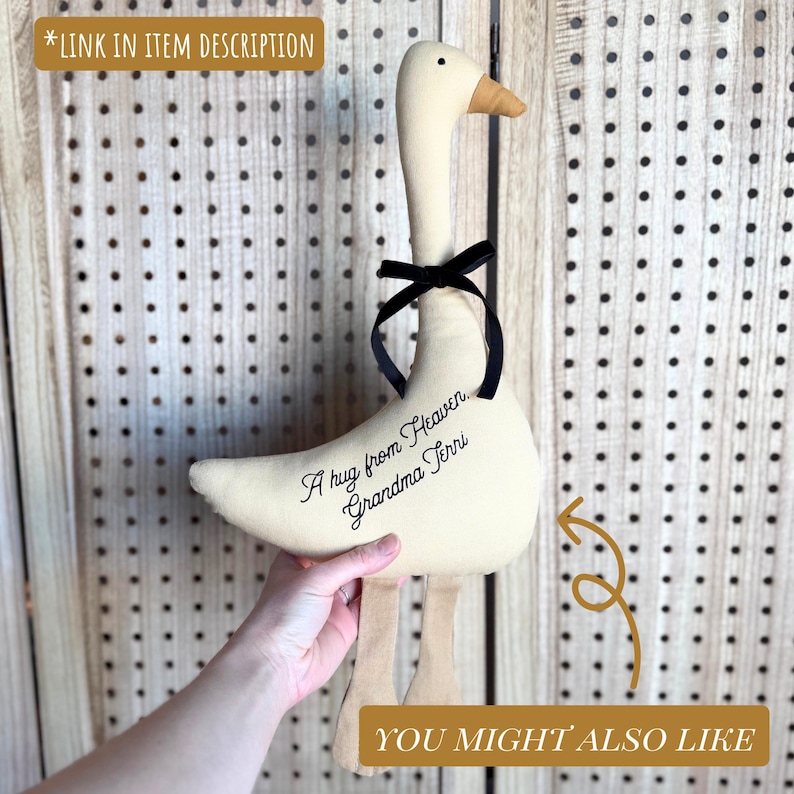 May include: A plush goose with a cream-colored body, a black ribbon, and tan legs and beak. The text "A hug from Heaven, Grandma Terri" is embroidered on the goose. The background is a light-colored pegboard.