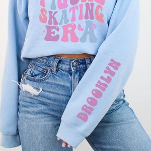 K&ouml;nnte beinhalten: Drei verk&uuml;rzte Sweatshirts in Pink, Hellblau und Dunkelblau mit dem Text "IN MY FIGURE SKATING ERA" in einer Retro-Schrift. Die &Auml;rmel jedes Sweatshirts haben das Wort "BROOKLYN" in pinker Schrift aufgedruckt.