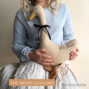May include: A beige plush goose toy with a yellow beak and black bow. The goose has the name "Cora Jeanette" embroidered on its side. The image also includes the text "YOU MIGHT ALSO LIKE" and "*LINK IN ITEM DESCRIPTION".