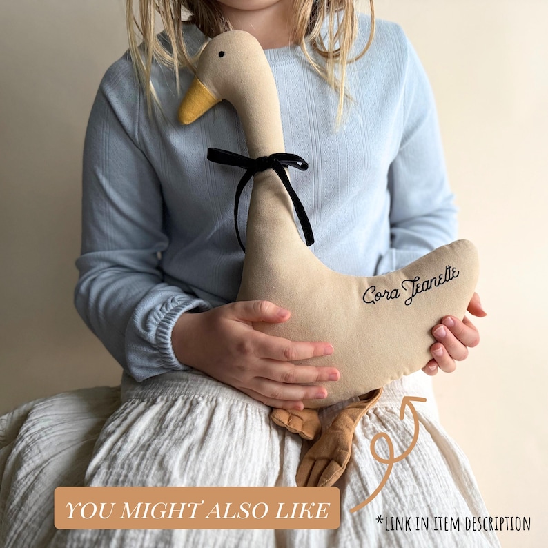 May include: A beige plush goose toy with a yellow beak and black bow. The goose has the name "Cora Jeanette" embroidered on its side. The image also includes the text "YOU MIGHT ALSO LIKE" and "*LINK IN ITEM DESCRIPTION".