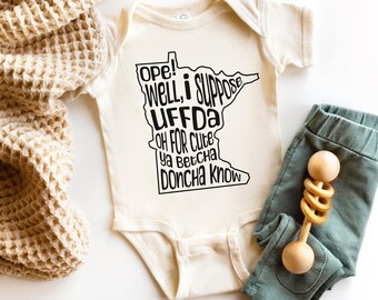 May include: Cream-colored baby onesie with black text in the shape of the state of Minnesota. The text reads "Ope! Well, I Suppose UFFDA Oh For Cute Ya Betcha Doncha Know." Beside the onesie are teal baby pants and a wooden rattle.
