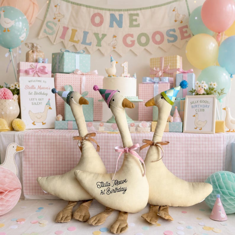 Puede incluir: Tres peluches de ganso de tela color crema con sombreros de fiesta coloridos y cintas alrededor del cuello. El ganso central tiene un bordado negro que dice Stella Maeve 1st Birthday. Cada ganso tiene patas naranjas y un cuello largo.