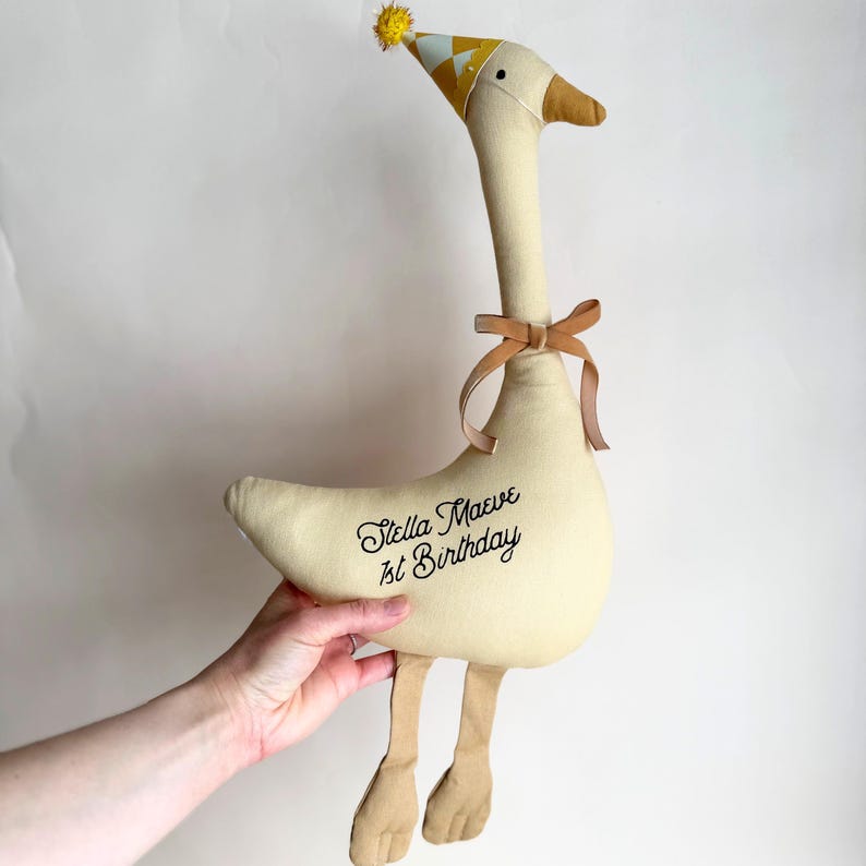 Puede incluir: Un peluche de tela color crema con forma de ganso que lleva un gorro de fiesta beige con un pomp&oacute;n amarillo, un lazo beige alrededor del cuello y el texto bordado en negro "Stella Maeve 1st Birthday" en el lateral, con dos patas de tela colgantes.