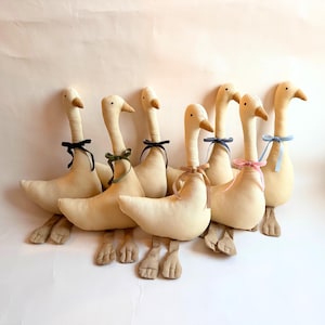 May include: A group of seven plush geese in a row, each with a cream-colored body, long neck, and brown beak. Each goose has a different colored ribbon tied around its neck, including black, green, and pink.