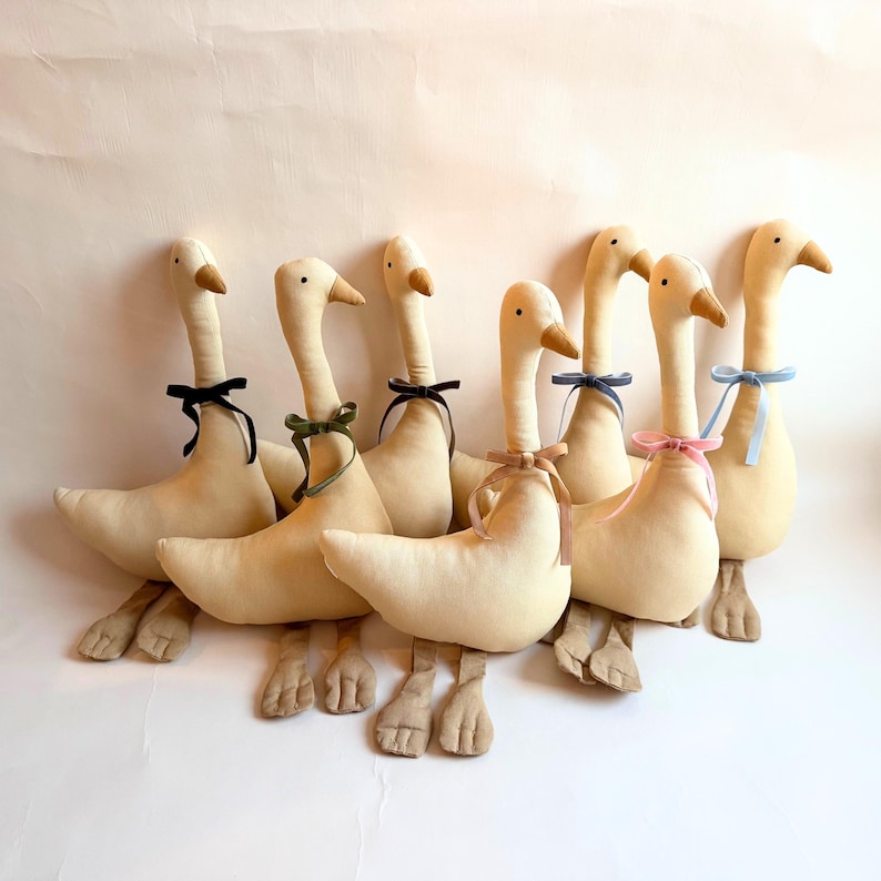 May include: A group of seven plush geese in a row, each with a cream-colored body, long neck, and brown beak. Each goose has a different colored ribbon tied around its neck, including black, green, and pink.