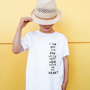 以下が含まれることがあります： 「I've got the Joy Joy Joy Joy down in my HEART」の文字が黒で縦にプリントされた白いTシャツ。黄色い背景を背景に、そのTシャツと茶色のバンドが付いた麦わら帽子を身に着けている人。