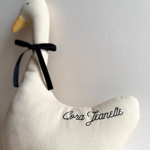 May include: A stuffed goose toy with a cream-colored body, a yellow beak, and a black velvet ribbon tied around its neck. The name "Cora Jeanette" is embroidered in black script on the goose's body. The toy is made of soft fabric.