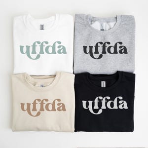 May include: Four sweatshirts in white, gray, beige, and black, each with the word "uffda" in a different color. The white sweatshirt has the word in a muted teal, the gray in black, the beige in brown, and the black in white. The sweatshirts are laid out on a white surface.