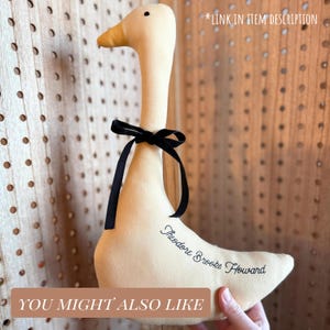 May include: A plush, cream-colored goose with a black ribbon tied around its neck. The goose has a yellow beak and the text "Theodore Brooks Howard" is embroidered on its side. The words "YOU MIGHT ALSO LIKE" and "*LINK IN ITEM DESCRIPTION" are also visible.