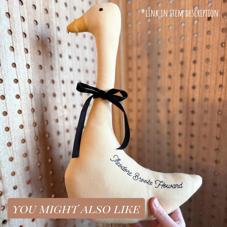 May include: A plush, cream-colored goose with a black ribbon tied around its neck. The goose has a yellow beak and the text "Theodore Brooks Howard" is embroidered on its side. The words "YOU MIGHT ALSO LIKE" and "*LINK IN ITEM DESCRIPTION" are also visible.