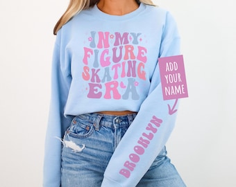 May include: Light blue cropped sweatshirt with the text "In My Figure Skating Era" in pink, blue, and white lettering. The sweatshirt has a long sleeve with the text "Brooklyn" in pink lettering. A pink rectangle with the text "Add Your Name" is in the upper right corner of the image.