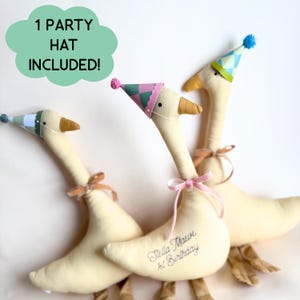 Puede incluir: Tres gansos de peluche color crema con peque&ntilde;os sombreros de fiesta c&oacute;nicos con pompones. Un ganso presenta el texto bordado "Stella Maeve 1st Birthday" y una cinta rosa. Un globo de texto verde indica "1 PARTY HAT INCLUDED!"