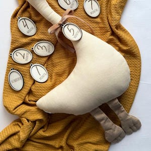 May include: A plush goose toy with a brown beak and feet, and a ribbon around its neck holding a wooden tag. The tag displays the number "5". Several wooden tags with months 1-12 are scattered around the goose on a mustard-colored blanket. The text "PLUSH MONTHLY MILESTONE TRACKER" is at the bottom.