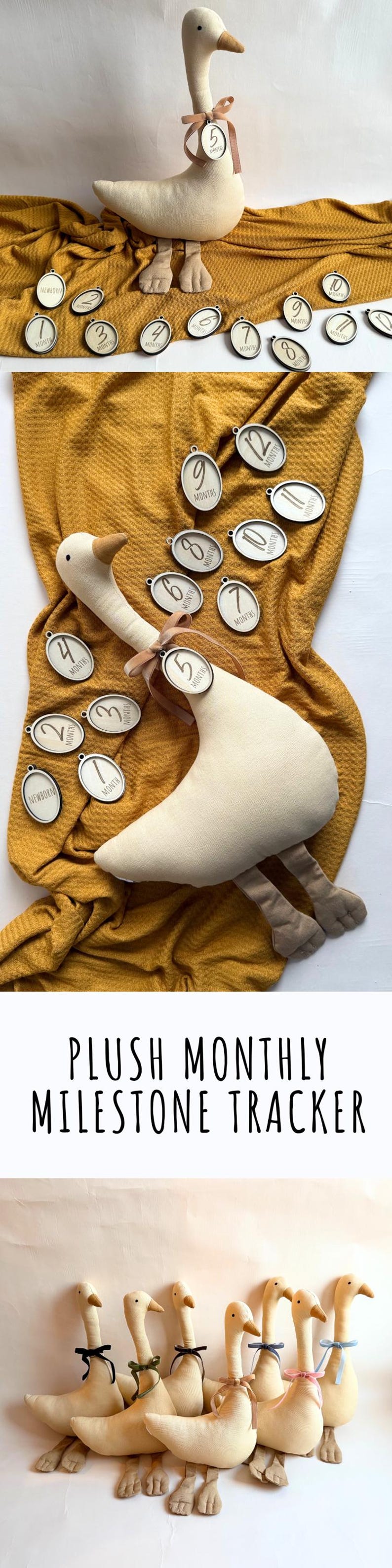 May include: A plush goose toy with a brown beak and feet, and a ribbon around its neck holding a wooden tag. The tag displays the number "5". Several wooden tags with months 1-12 are scattered around the goose on a mustard-colored blanket. The text "PLUSH MONTHLY MILESTONE TRACKER" is at the bottom.