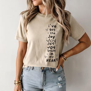 May include: A beige t-shirt with the text "I've got the Joy Joy Joy Joy Down in my HEART" in black script. Small gray hearts are scattered around the text. The shirt is paired with light blue denim shorts.