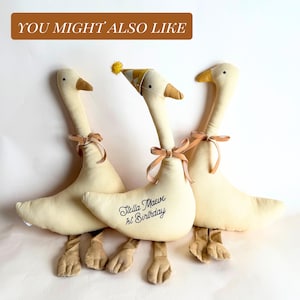 May include: Three plush goose toys in cream with brown beaks and feet. Each goose has a tan ribbon tied around its neck. One goose wears a party hat and has the text "Stella Maeve 1st Birthday" on its body.