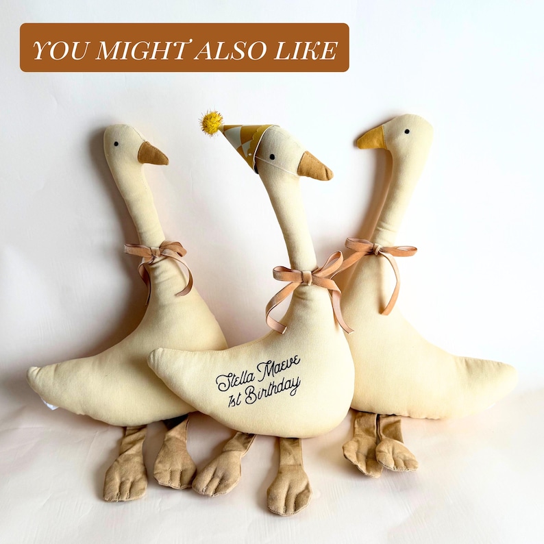 May include: Three plush goose toys in cream with brown beaks and feet. Each goose has a tan ribbon tied around its neck. One goose wears a party hat and has the text "Stella Maeve 1st Birthday" on its body.