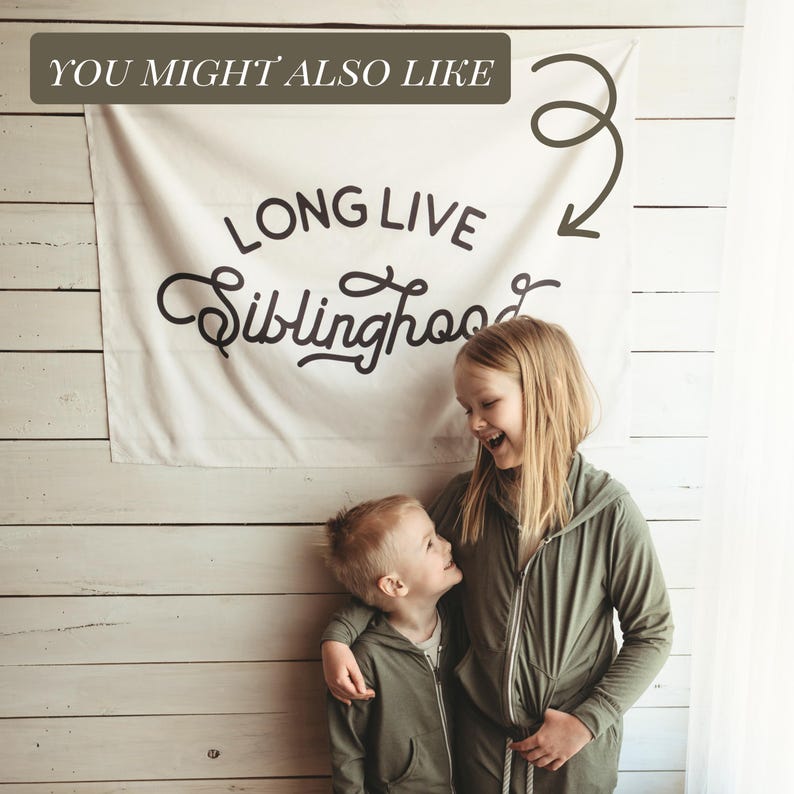 May include: A white banner with the text "LONG LIVE Siblinghood" in black script. Two children are wearing olive green zip-up hoodies. The banner is hanging on a white wooden wall.