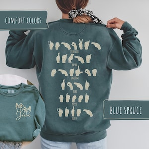 May include: A teal blue sweatshirt with a graphic of sign language hand gestures and the words "Jehovah", "Kapha", "Shalom", "Rohs", "Nissi", and "Jireh" printed on the back. The sweatshirt also has the words "Comfort Colors" and "Blue Spruce" printed on it.