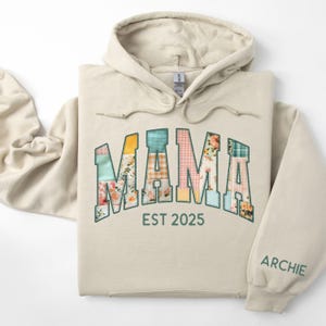 May include: A beige hoodie with the word "MAMA" in patchwork letters, featuring floral and plaid patterns. The text "EST 2025" is below the word "MAMA". The name "ARCHIE" is printed on the sleeve.