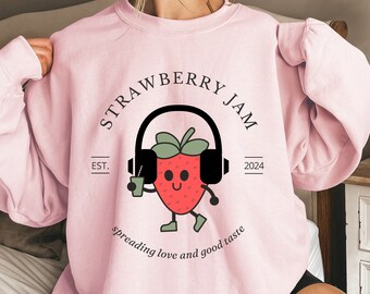 Könnte beinhalten: Rosafarbener Pullover mit einem Motiv einer Cartoon-Erdbeere, die Kopfhörer trägt und ein Getränk hält. Der Text "Strawberry Jam" ist in einem Kreis um die Erdbeere geschrieben. Der Text "Est. 2024" ist unter der Erdbeere geschrieben. Der Text "spreading love and good taste" ist unter dem Kreis geschrieben.