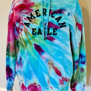 May include: A tie-dye hoodie with the words "American Eagle" in black lettering on the front. The hoodie is a mix of blue, green, pink, and purple.