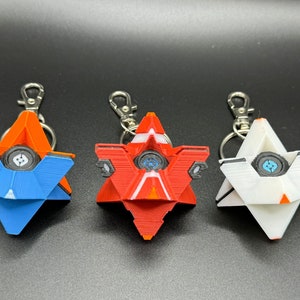 May include: Three 3D printed keychains shaped like a star with a round eye in the centre. The keychains are blue, red and white.