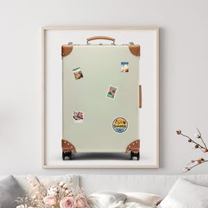 Op de afbeelding: Een crèmekleurige koffer in vintage-stijl met bruine lederen hoekverstevigingen en een handvat aan de bovenkant. De koffer is voorzien van vier kleine reisstickers, waaronder een ronde sticker met de tekst Bahamas, in een houten lijst.