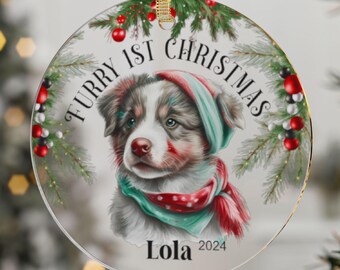 May include: A round glass Christmas ornament with a white background and a cute cartoon dog wearing a red and green hat and scarf. The text "Furry 1st Christmas" is written in black lettering around the dog. The dog's name "Lola" and the year "2024" are written below the dog.