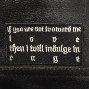 Puede incluir: Un parche rectangular negro bordado cosido sobre una superficie de cuero negro. El parche presenta un texto blanco de estilo gótico que dice: If you are not toward me love then i will indulge in rage, con las palabras love y rage espaciadas.