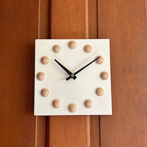 May include: A white square clock with black hands and wooden hour markers. The clock is mounted on a wooden wall.
