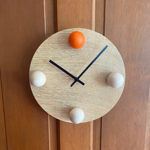 Puede incluir: Un reloj redondo de madera con manecillas negras y tres pomos de madera. El reloj tiene un pomo naranja en la parte superior.