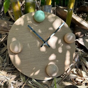 Puede incluir: Un reloj redondo de madera con un acabado marr&oacute;n claro. El reloj tiene una esfera de metal negro con manecillas plateadas y cuatro pomos redondos, uno verde, uno marr&oacute;n claro y dos marr&oacute;n oscuro.