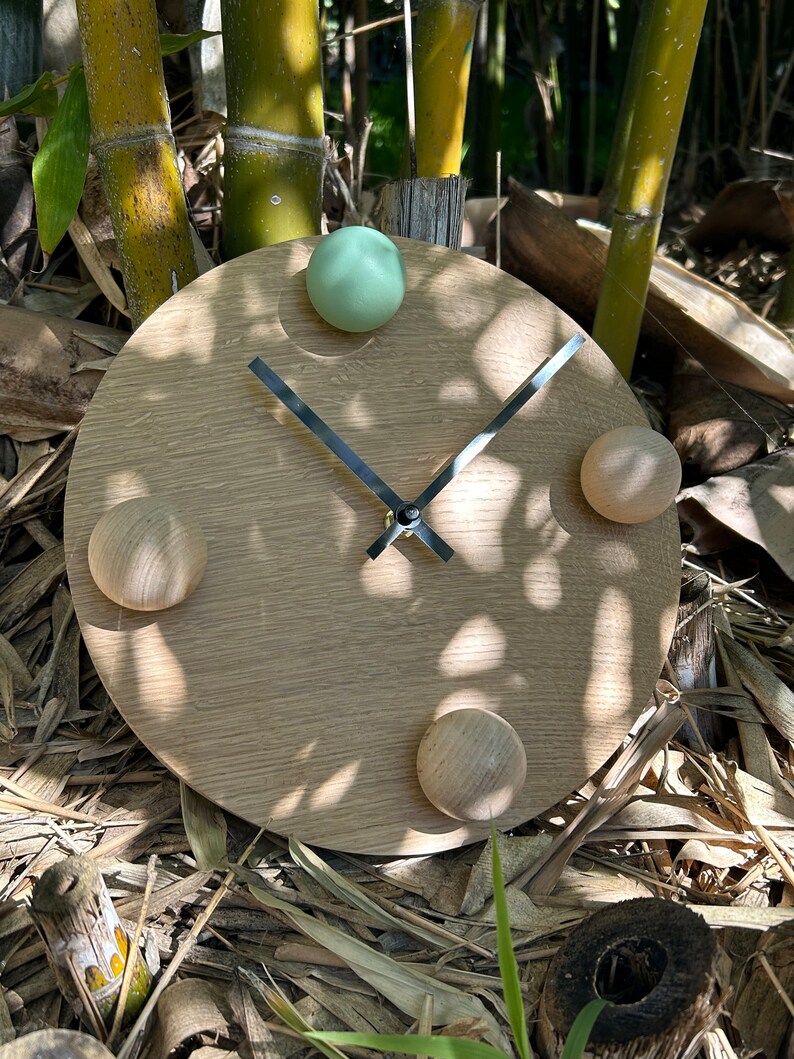 Puede incluir: Un reloj redondo de madera con un acabado marr&oacute;n claro. El reloj tiene una esfera de metal negro con manecillas plateadas y cuatro pomos redondos, uno verde, uno marr&oacute;n claro y dos marr&oacute;n oscuro.
