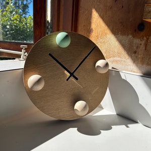 Puede incluir: Un reloj de madera con una esfera de color marr&oacute;n claro y agujas negras. El reloj tiene cuatro pomos redondos, uno verde, uno marr&oacute;n claro y dos marr&oacute;n oscuro.