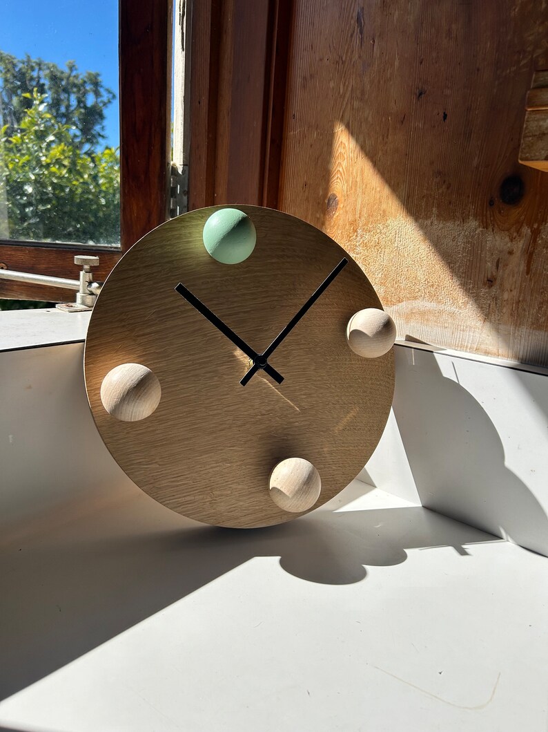 Puede incluir: Un reloj de madera con una esfera de color marr&oacute;n claro y agujas negras. El reloj tiene cuatro pomos redondos, uno verde, uno marr&oacute;n claro y dos marr&oacute;n oscuro.